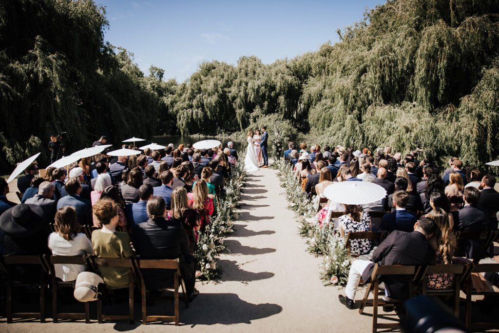 An outdoor wedding ceremony with guests seated on either side of a flower-lined aisle, facing a couple standing with an officiant under clear skies and surrounded by trees.