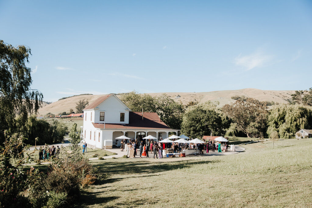 A group of people gather outside a white two-story house with a red roof, set in a rural landscape with hills and trees under a clear sky.