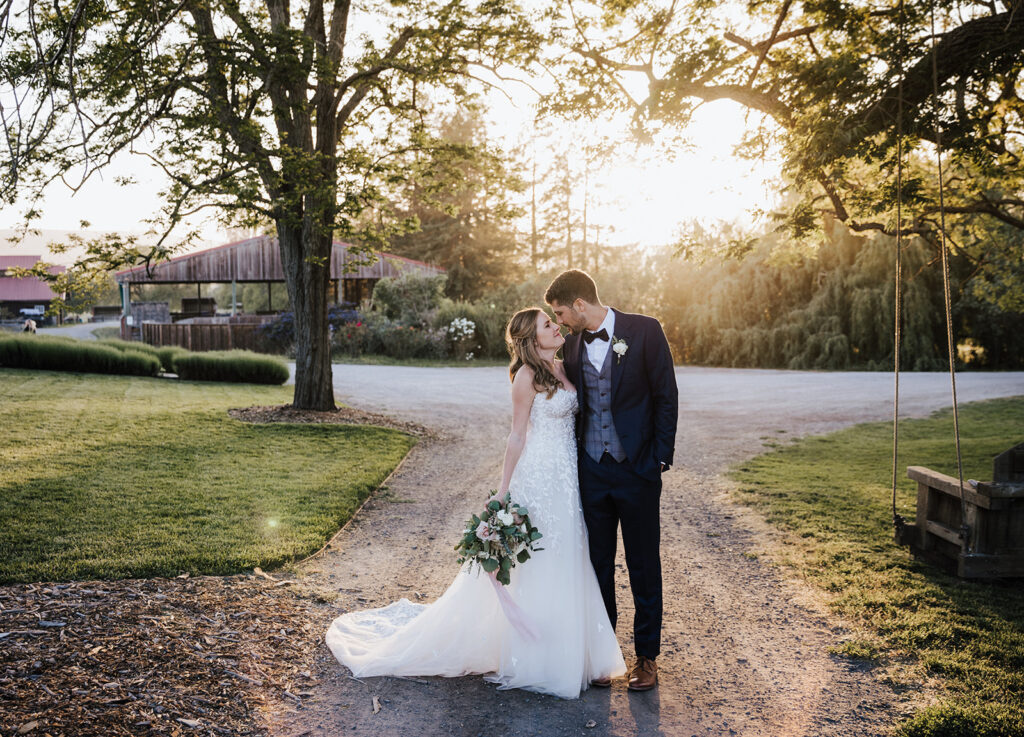 A bride and groom stand close together on a tree-lined path at sunset, with the bride holding a bouquet and the groom leaning in for a kiss.