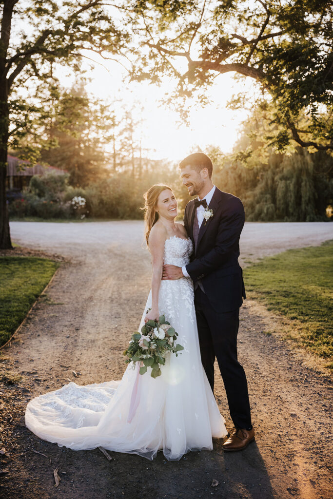A bride and groom stand together outdoors at sunset, smiling, with the bride holding a bouquet and wearing a white dress, and the groom in a dark suit.