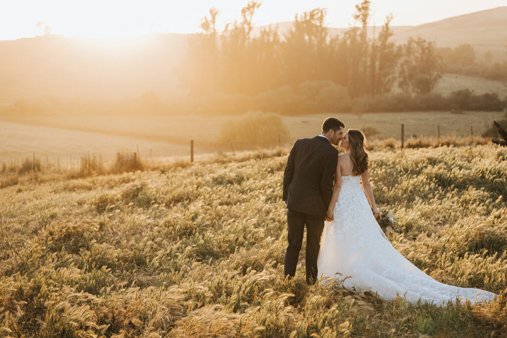 A bride and groom stand together in a grassy field at sunset, facing away from the camera, with trees and hills in the background.