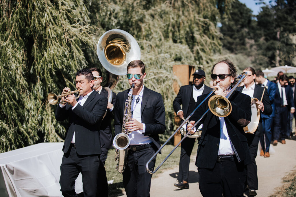 A brass band in black suits plays instruments, including trumpet, saxophone, trombone, and sousaphone, while marching outdoors along a tree-lined path.