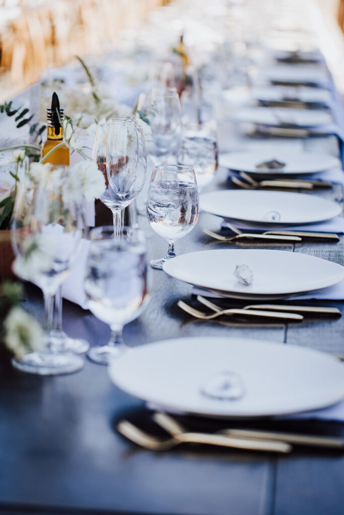 A long, elegant dining table set with white plates, glasses, cutlery, and floral centerpieces, ready for guests.