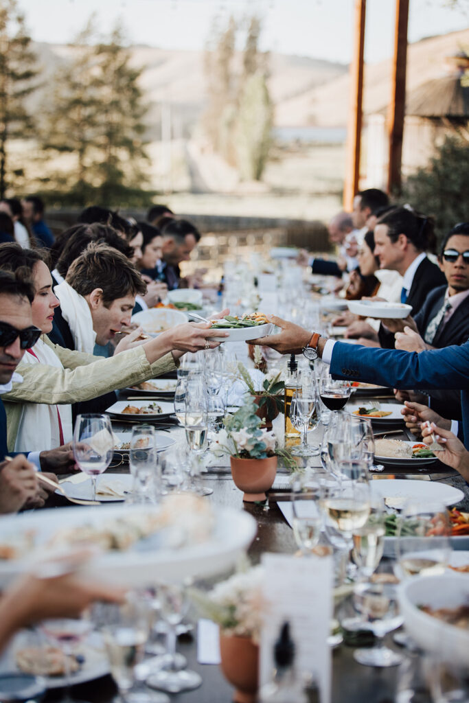 People seated at a long outdoor table share food and drinks during a formal gathering, with glassware and plated dishes visible.