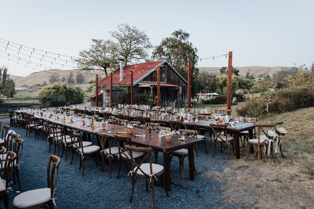 Long wooden tables with chairs are set up outdoors for an event near a rustic barn, with string lights overhead and countryside scenery in the background.
