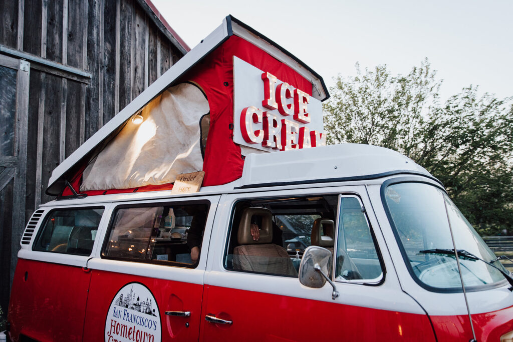 A red and white vintage van with a pop-up roof and large "ICE CREAM" sign is parked next to a rustic wooden building.