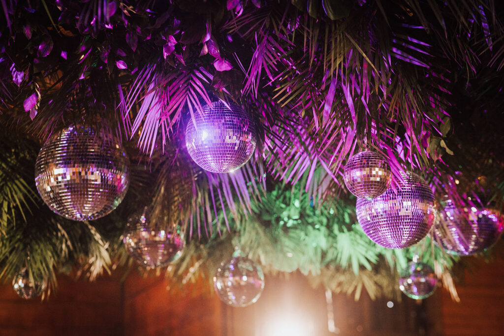 Disco balls hang from a ceiling decorated with green palm leaves and lit by purple and pink lights.