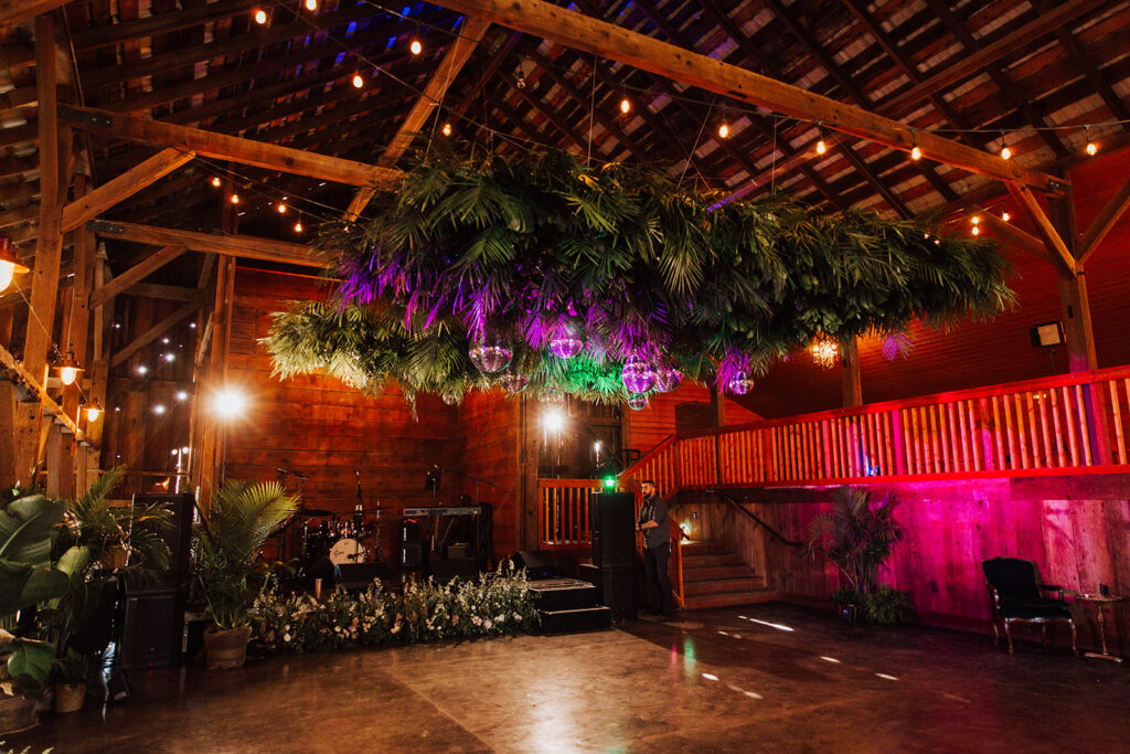 Large rustic barn interior with wooden beams, string lights, and greenery suspended from the ceiling; stage area and plants arranged along the floor.