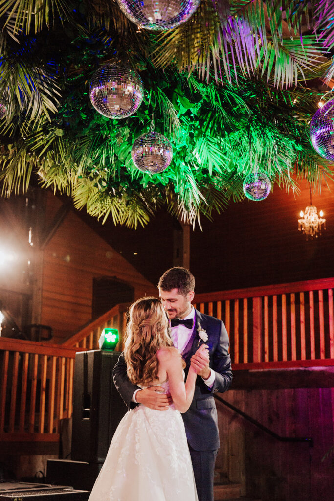 A bride and groom dance together under hanging greenery and disco balls at an indoor wedding venue.