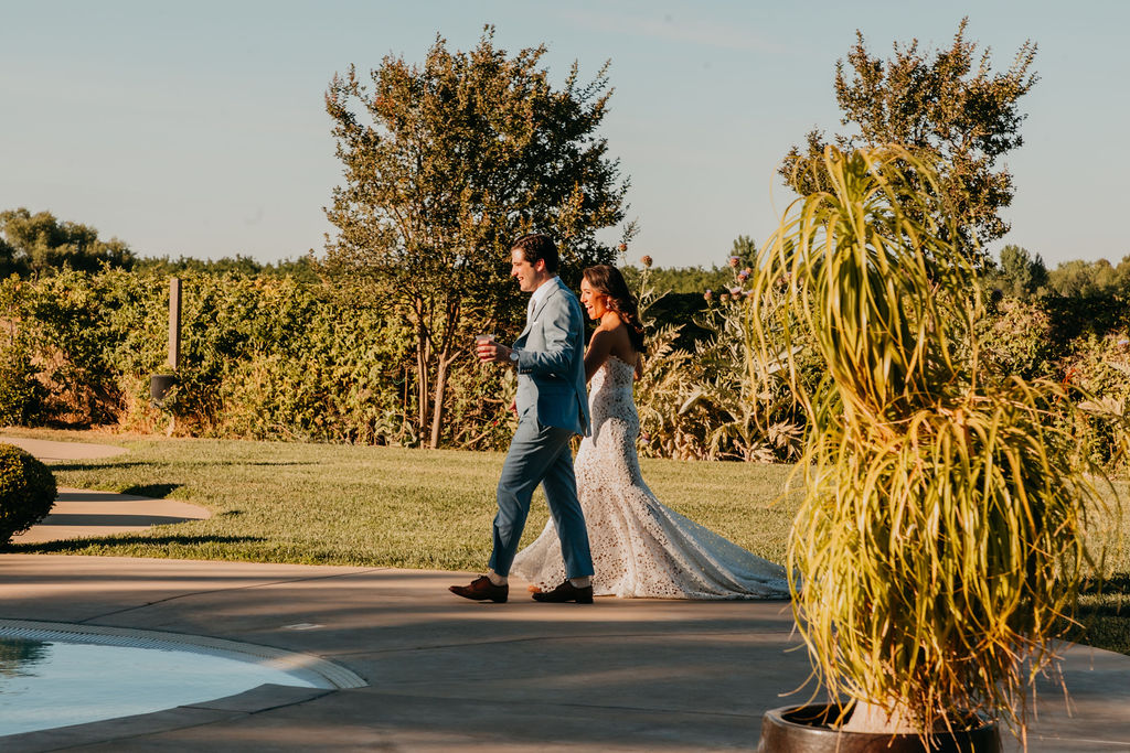A bride in a white dress and a groom in a blue suit walk side by side along a path near a pool, surrounded by greenery and trees.