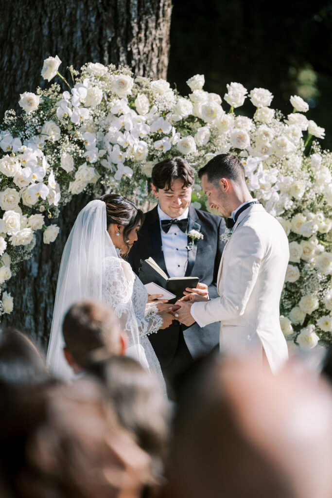 A bride and groom stand before an officiant, exchanging rings in an outdoor wedding ceremony with a white floral arch in the background.