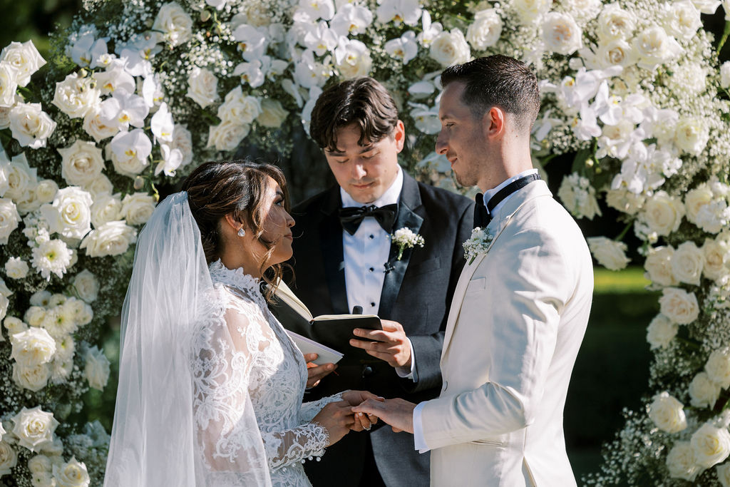 A bride and groom stand facing each other, holding hands, as an officiant reads from a book during an outdoor wedding ceremony in front of a floral arch.