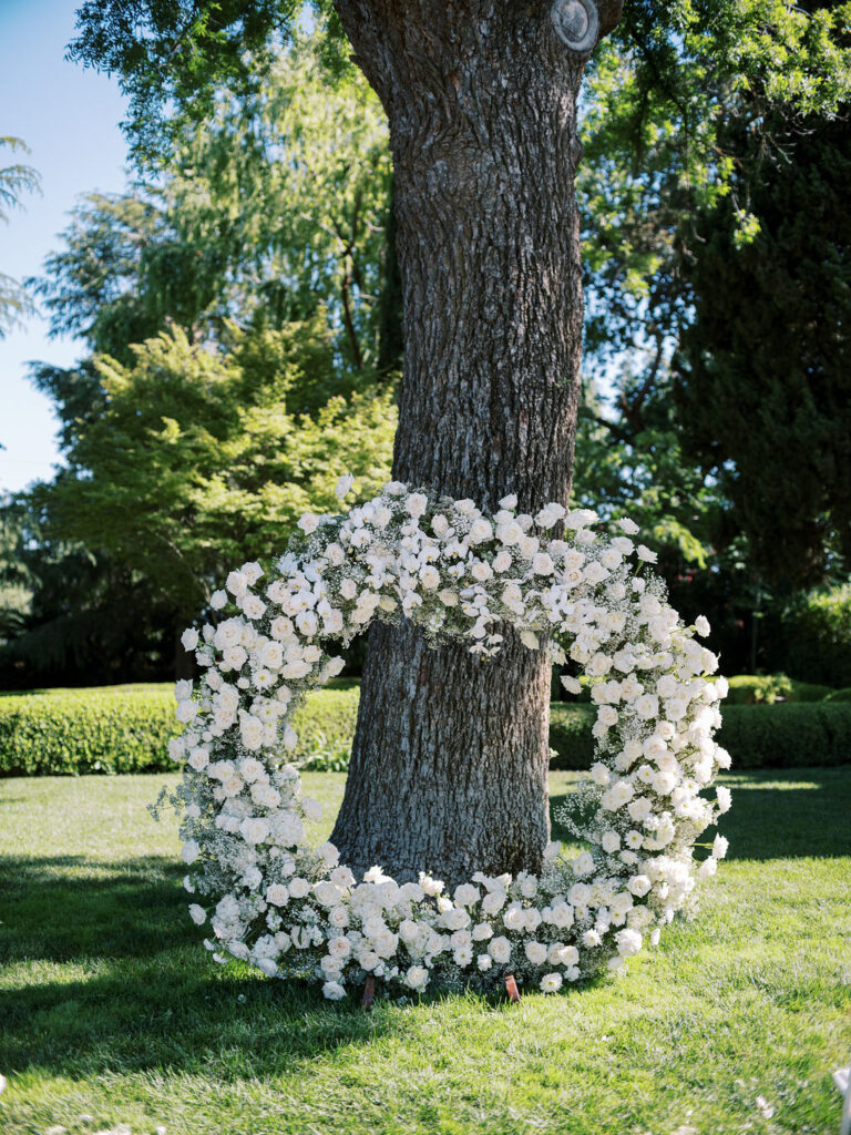Large circular floral arrangement of white flowers stands in front of a tree in a green garden setting on a sunny day.