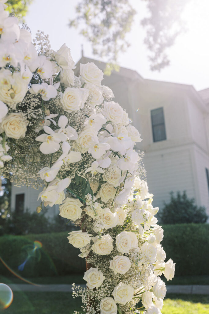 A floral arch made of white roses, orchids, and baby's breath stands outdoors in front of a white house on a sunny day.