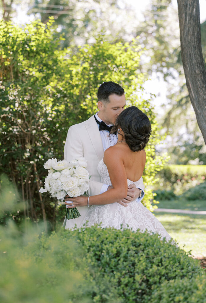 A bride and groom embrace outdoors; the groom in a white tuxedo kisses the bride, who holds a bouquet of white flowers. Lush greenery surrounds them.