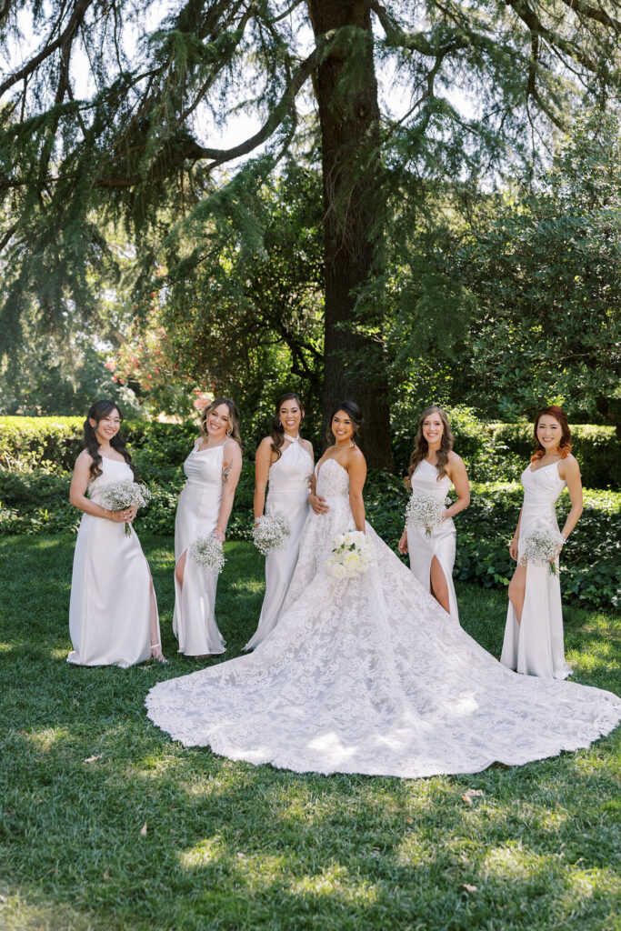 A bride in a lace gown stands with five bridesmaids in white dresses holding bouquets, posing outdoors on grass with trees and greenery in the background.
