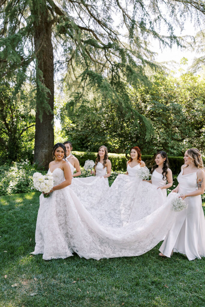 A bride in a lace gown holds a bouquet as four bridesmaids in white dresses help arrange her train outdoors under tall trees.