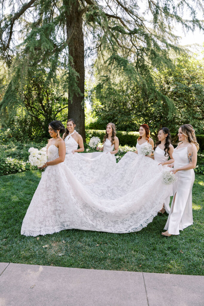 A bride in a white lace gown stands outdoors as four bridesmaids in white dresses help arrange her train on a grassy area with trees in the background.