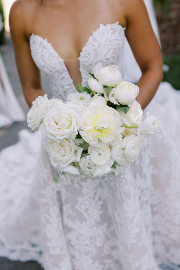 A bride in a strapless lace wedding gown holds a bouquet of white flowers, including roses and peonies.