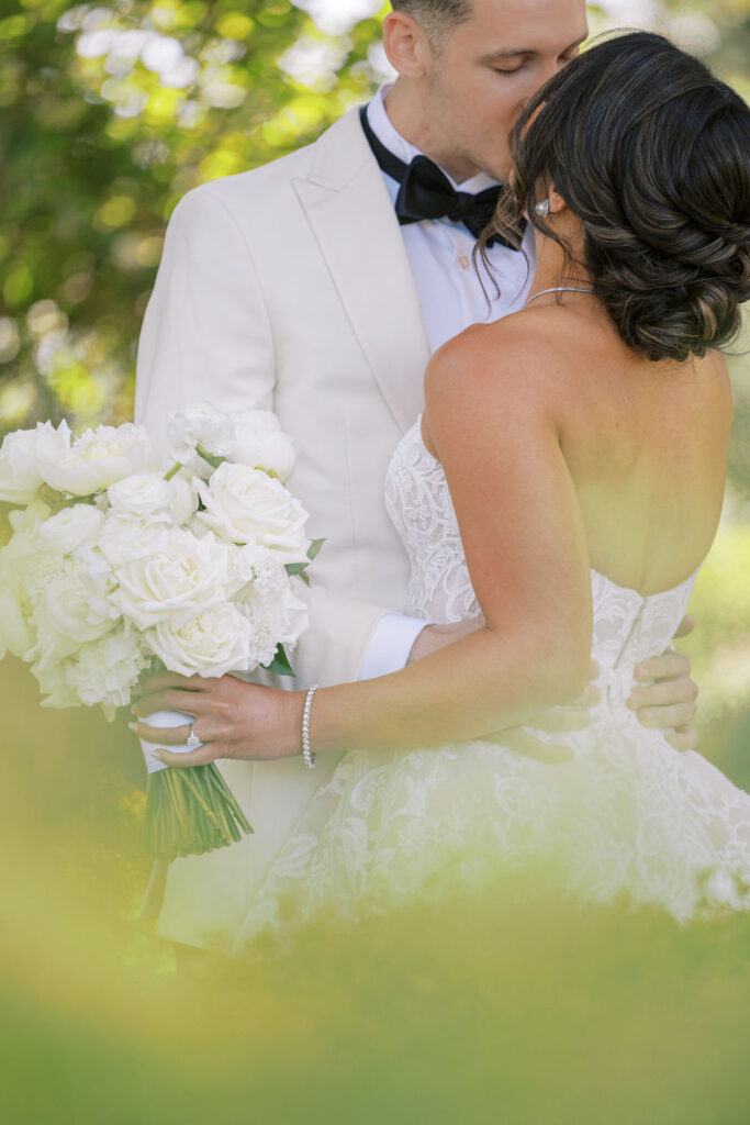 A bride in a strapless white gown holds a bouquet of white roses as she is embraced and kissed on the forehead by a groom in a white suit and black bow tie.