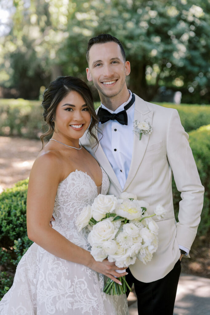 A bride in a white strapless dress and a groom in a white tuxedo jacket stand together outdoors, smiling and holding a bouquet of white flowers.