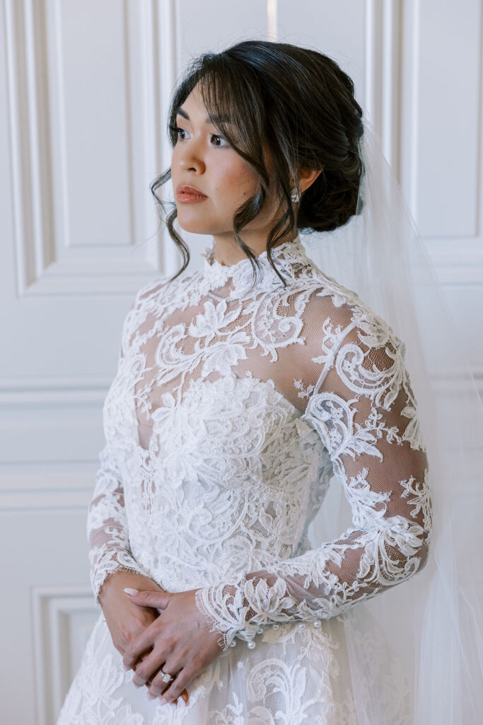 A woman in a long-sleeved, lace wedding gown with a high neckline and veil stands in front of a white paneled wall, looking off to the side.