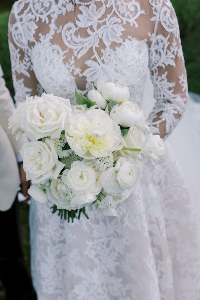 Bride in a white lace wedding dress holding a bouquet of white flowers, including roses and peonies, with greenery visible in the background.