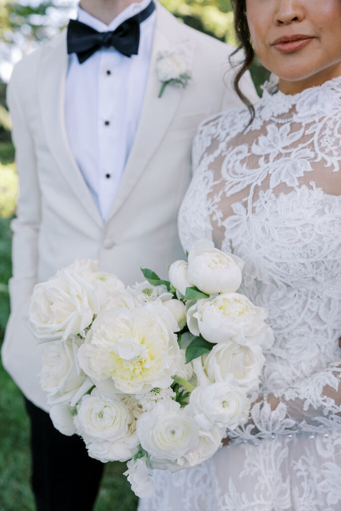 Bride in a lace wedding dress holds a white bouquet, standing next to groom in a white tuxedo jacket and black bow tie.