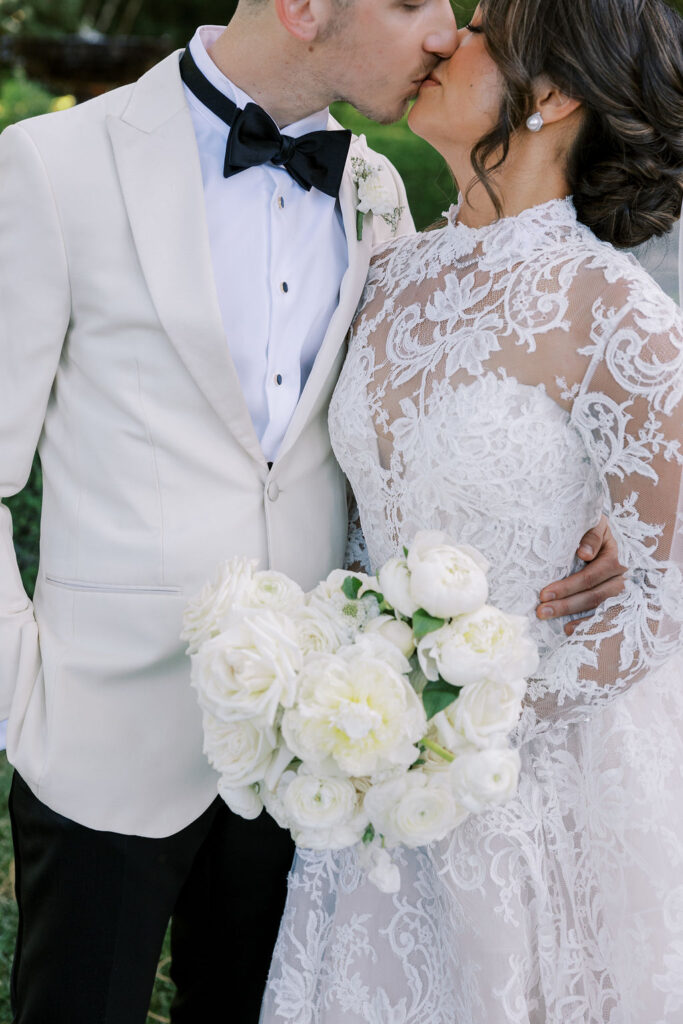 A bride and groom share a kiss. The bride wears a lace wedding dress and holds a bouquet of white flowers; the groom wears a white tuxedo jacket and black bow tie.