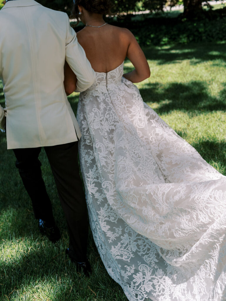 A couple is walking on grass, the person on the right is wearing a strapless white lace dress, and the person on the left is in a light-colored jacket and dark pants.