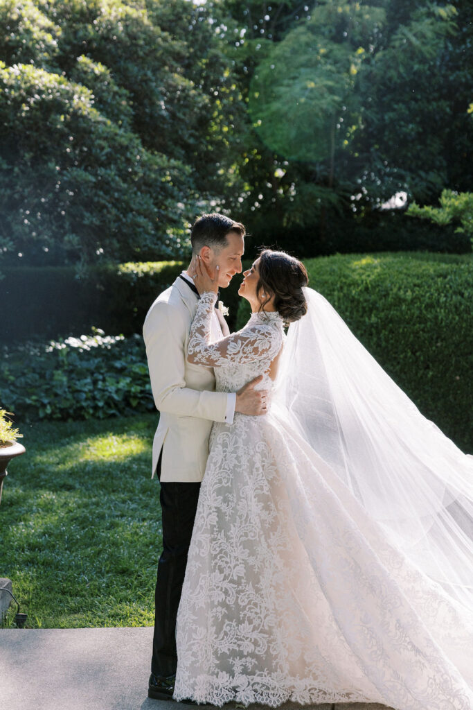 A bride and groom stand face-to-face outdoors, embracing and smiling, with greenery and sunlight in the background. The bride wears a long lace gown and veil; the groom wears a white jacket.