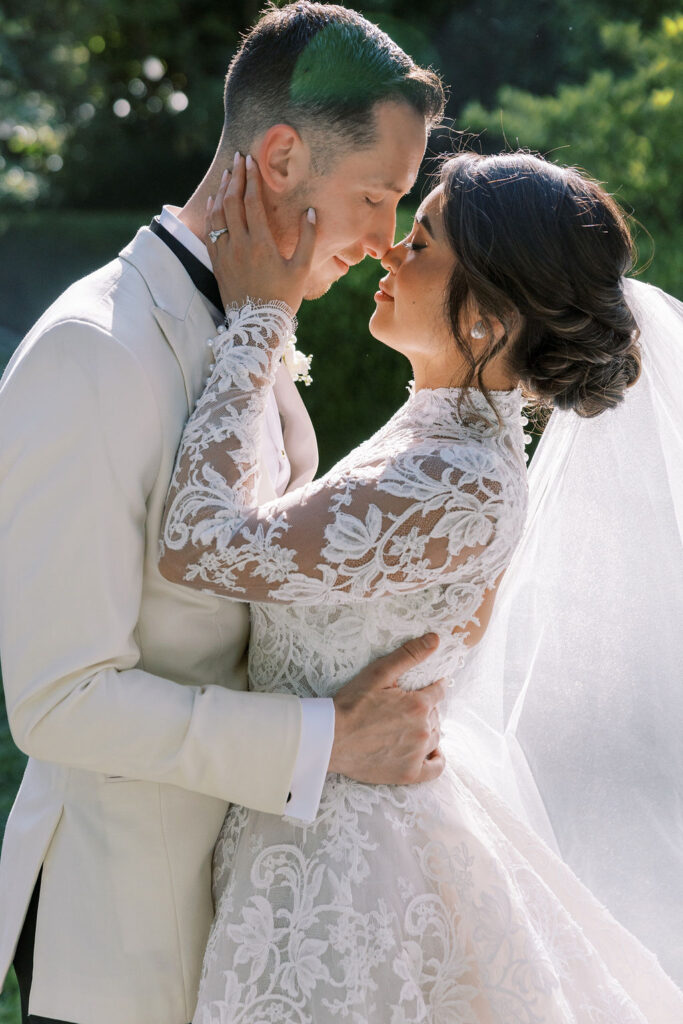 A bride and groom in formal wedding attire embrace outdoors, touching foreheads and closing their eyes, with sunlight highlighting the bride's lace dress and veil.