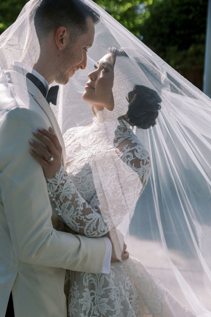 A bride and groom embrace under a lace veil outdoors, gazing at each other on their wedding day.