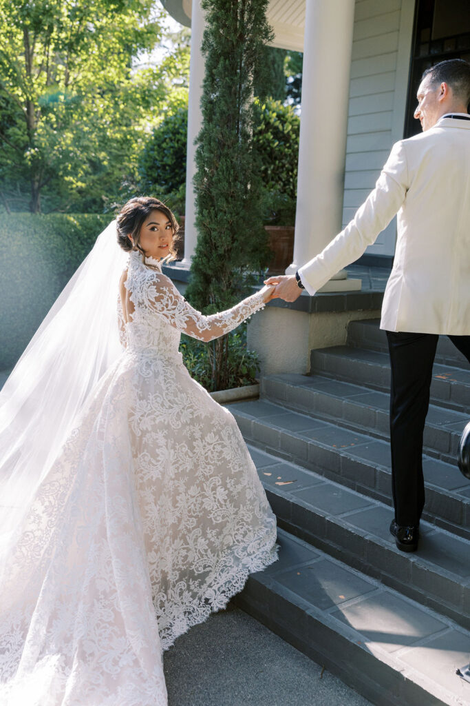 A bride in a lace wedding dress and veil holds hands with a groom in a white jacket as they walk up outdoor steps.
