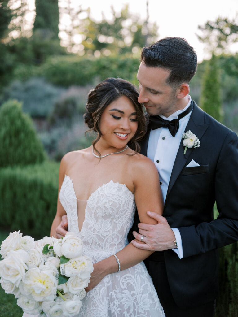 A bride in a lace wedding dress holding white roses stands with a groom in a black tuxedo, embracing outdoors in a garden.
