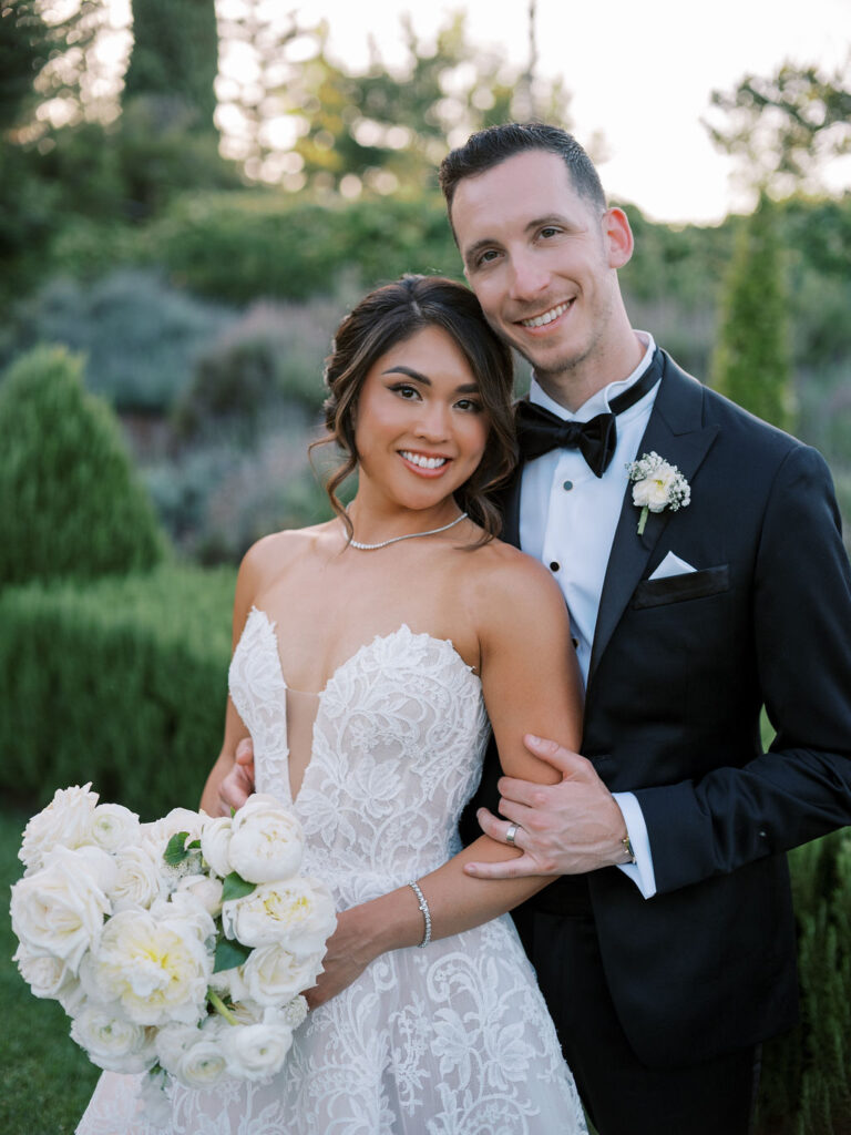 Bride and groom posing outdoors in formal wedding attire; bride holds a bouquet of white roses, and they are surrounded by greenery.