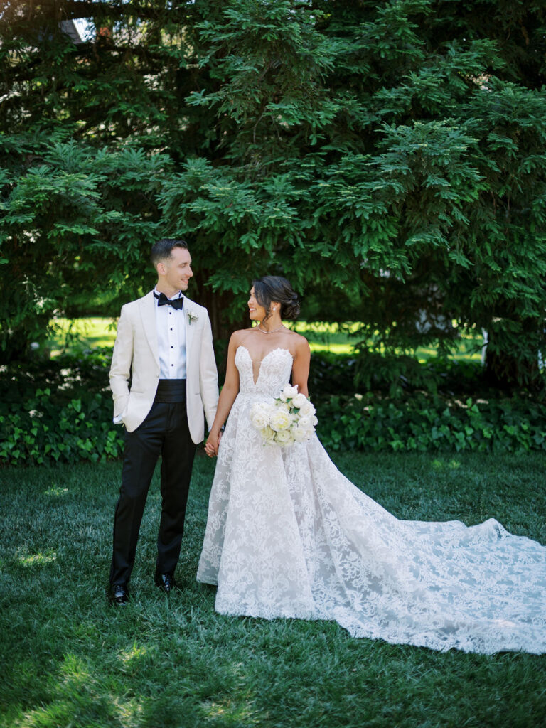 A bride in a white lace gown holds a bouquet and stands beside a groom in a light jacket and black pants; they are outdoors in front of green trees.