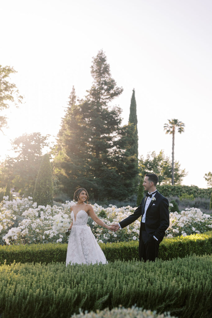 A bride and groom hold hands and face each other in a garden with blooming white flowers and tall trees in the background, dressed in wedding attire.
