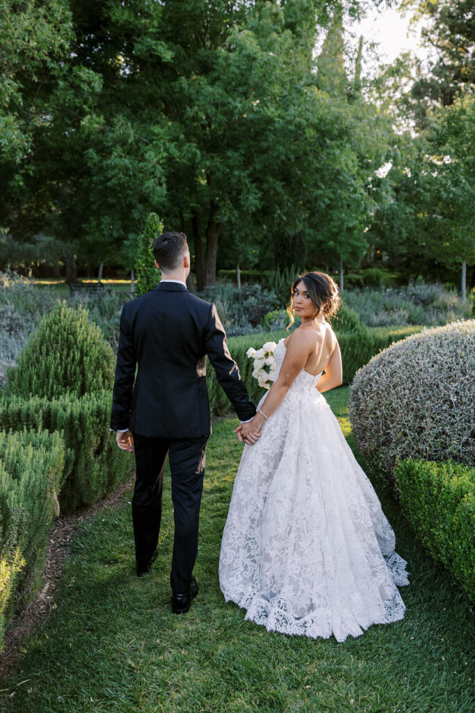 A bride and groom walk hand in hand through a garden, with the bride looking back over her shoulder.