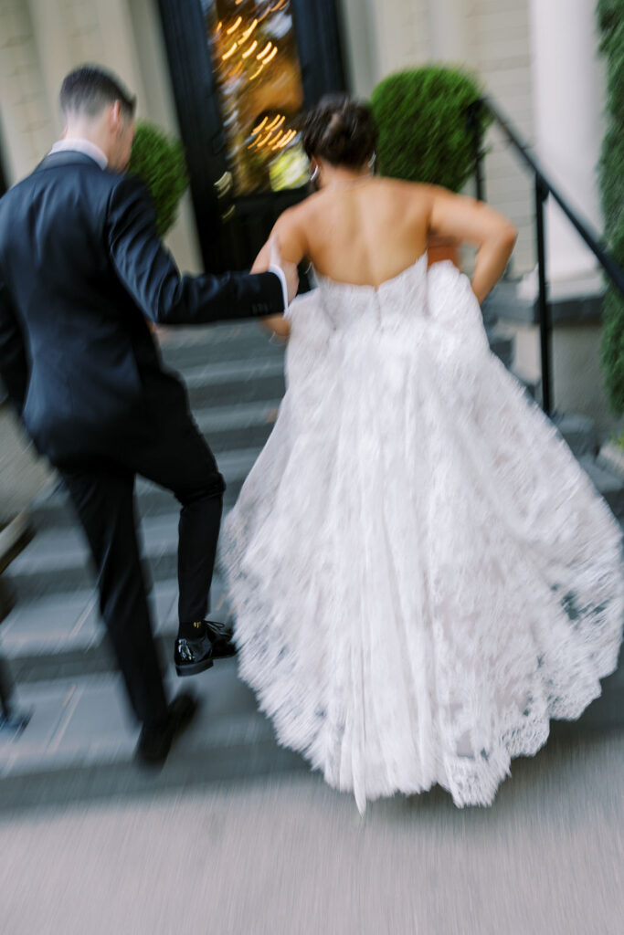 A bride in a white lace gown and a groom in a black suit walk up steps toward a building, holding hands. The image is slightly blurred, conveying movement.