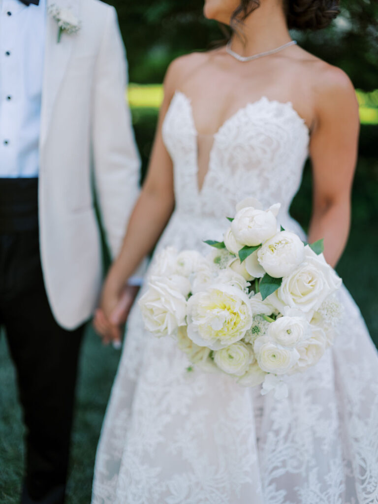 A bride in a white lace gown holding a white flower bouquet stands beside a groom in a white jacket and black pants. They are holding hands outdoors.