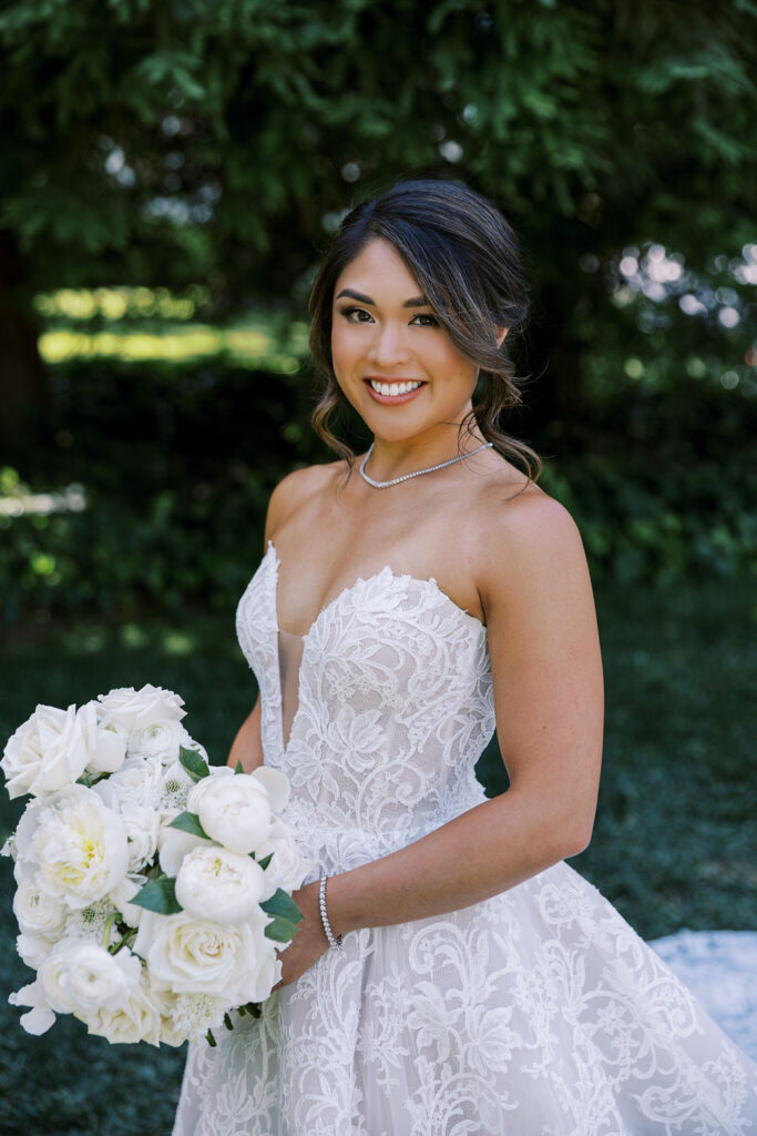 A woman in a strapless white lace wedding dress holds a bouquet of white flowers and smiles, standing outdoors with greenery in the background.