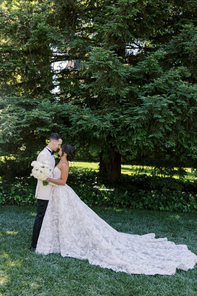 A bride and groom stand closely together on grass, surrounded by large green trees. The bride holds a bouquet and wears a long, flowing white dress with a train.