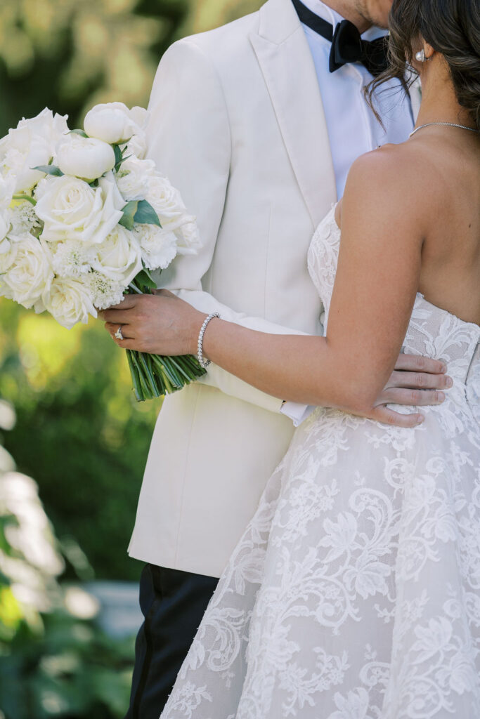 A bride in a lace gown holding a bouquet of white flowers stands with a groom in a white tuxedo jacket and black bow tie, embracing outdoors.