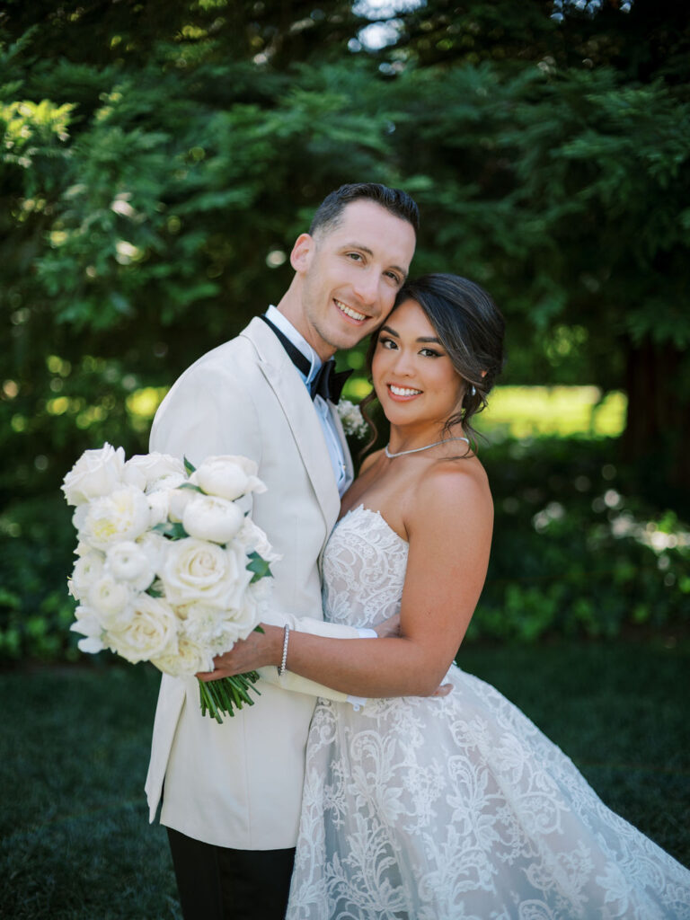 A bride and groom pose outdoors, embracing and smiling at the camera. The bride holds a bouquet of white roses and wears a strapless lace gown, while the groom wears a white tuxedo.