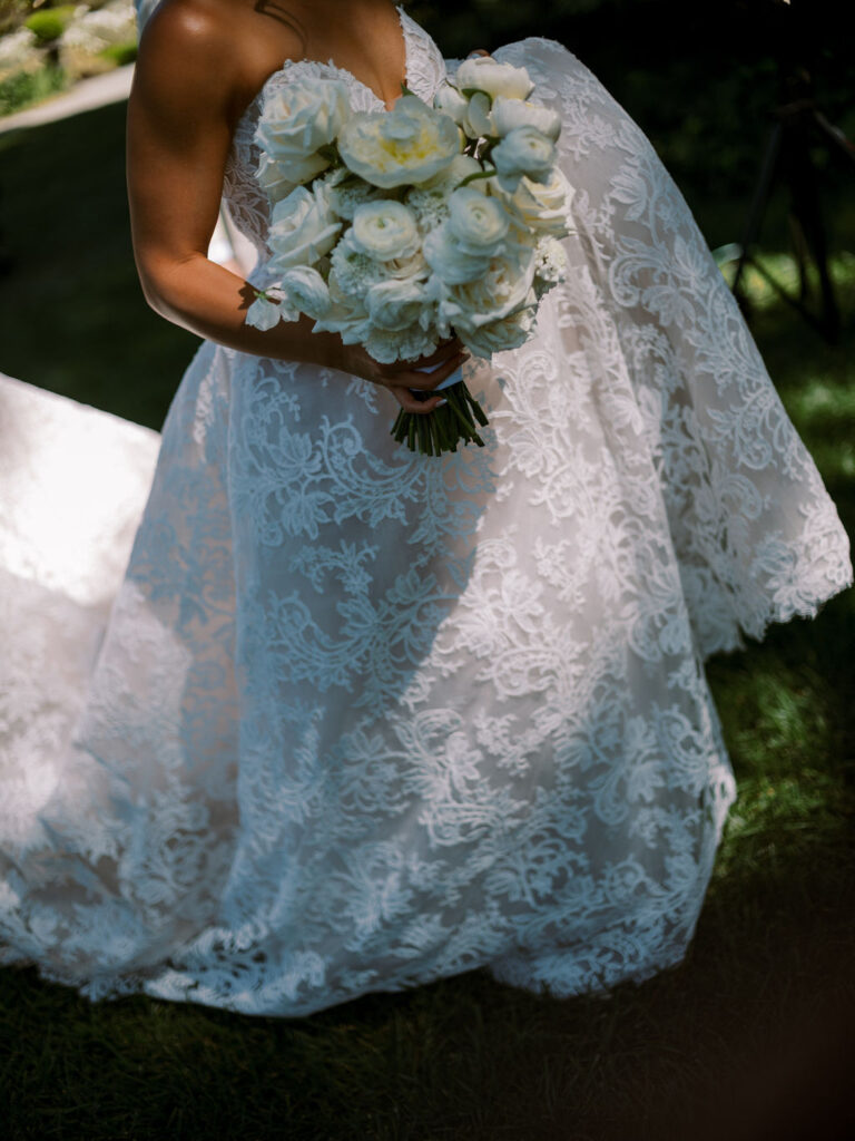 A person in a white lace wedding dress holds a bouquet of white flowers outdoors.