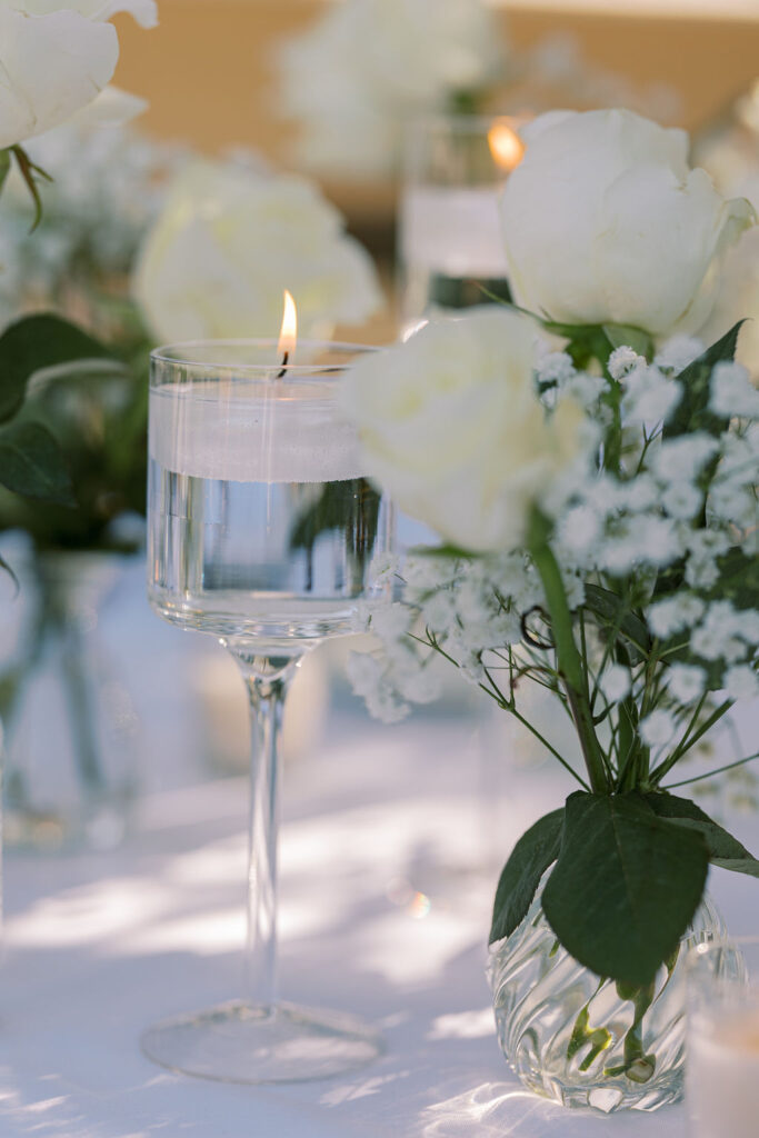 A floating candle in a glass of water is surrounded by white roses and baby's breath flowers in small glass vases on a white tablecloth.