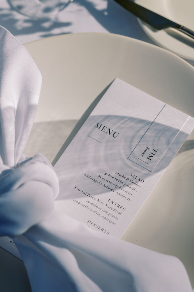 A white dinner plate with a neatly folded white napkin and a printed menu card listing salad, entrée, and dessert options.