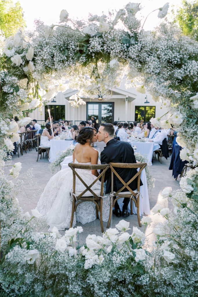 A bride and groom sit and kiss at their outdoor wedding reception, framed by a circular floral arrangement of white roses and baby's breath, with guests seated at tables in the background.