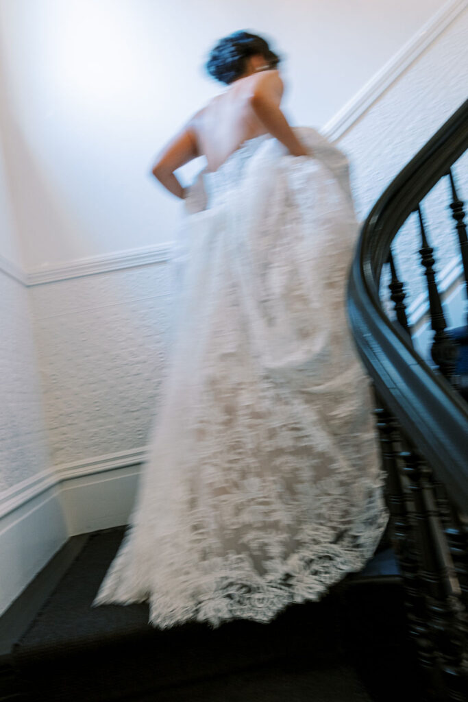 A person in a white lace wedding dress ascends a staircase, holding up the skirt. The image is slightly blurred, conveying motion.